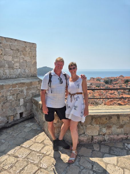 Paar steht auf einer Mauer mit Blick auf die Küste und Häuser im Hintergrund.