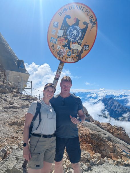 Ein Paar steht lächelnd vor einem Gipfelschild bei klarem Himmel und Berglandschaft.