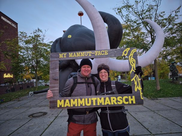 Zwei Personen posieren lachend vor einem großen, aufblasbaren Mammut.
