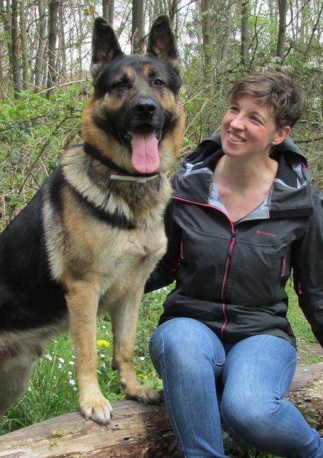 Eine lächelnde Frau sitzt neben einem großen Schäferhund im Wald.