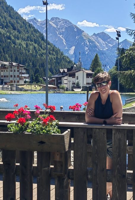 Frau mit Sonnenbrille steht an einem Holzbalkon, im Hintergrund Berge und ein See.