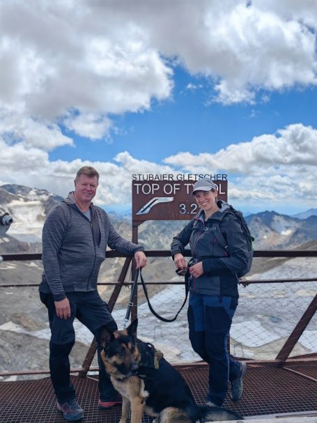 Zwei Personen stehen mit einem Hund am Aussichtspunkt „Top of the World“ mit Wolkenhimmel.